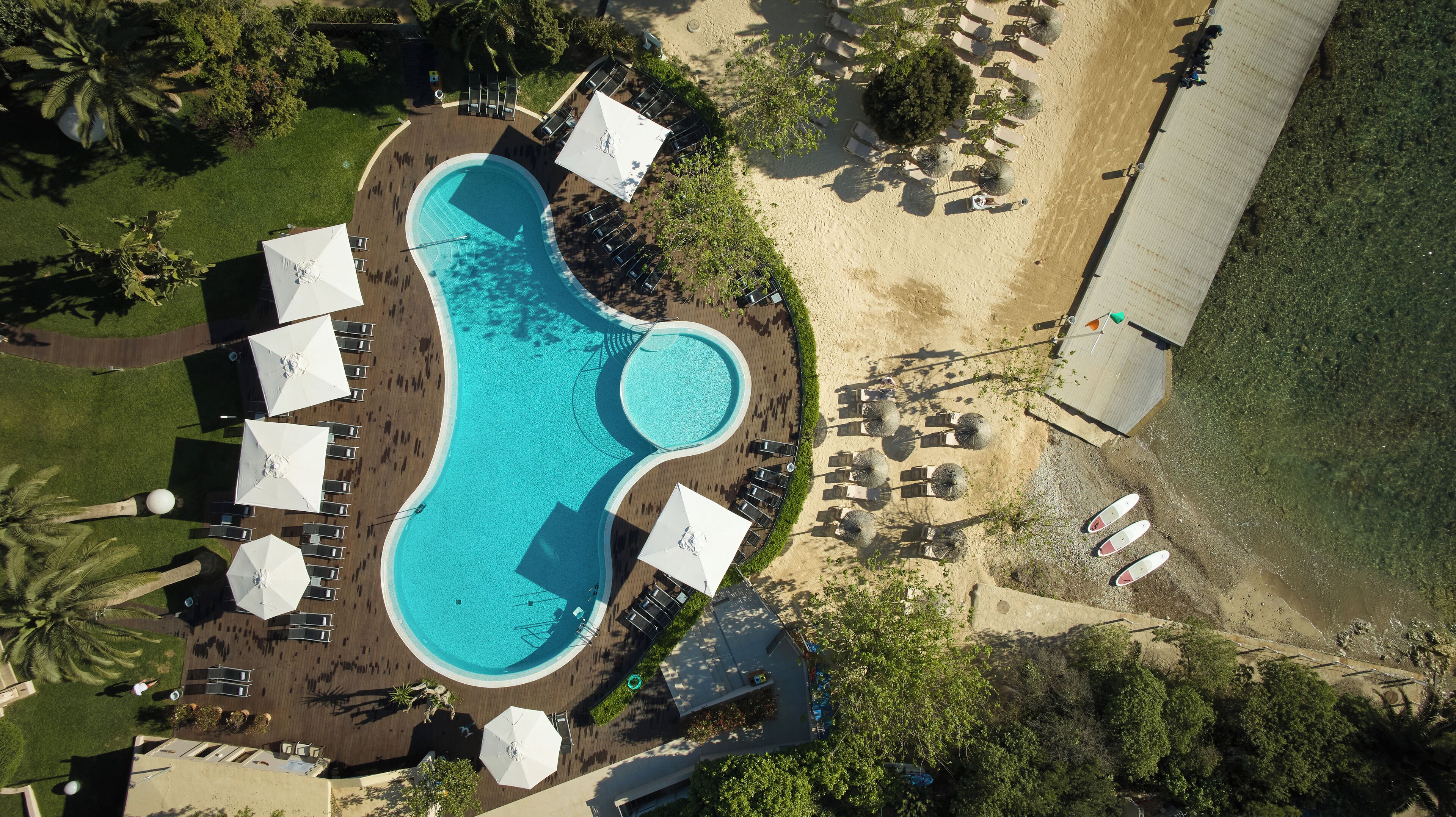 Vista aérea de un resort con una amplia piscina rodeada de tumbonas y sombrillas, una zona de playa arenosa y abundante vegetación.