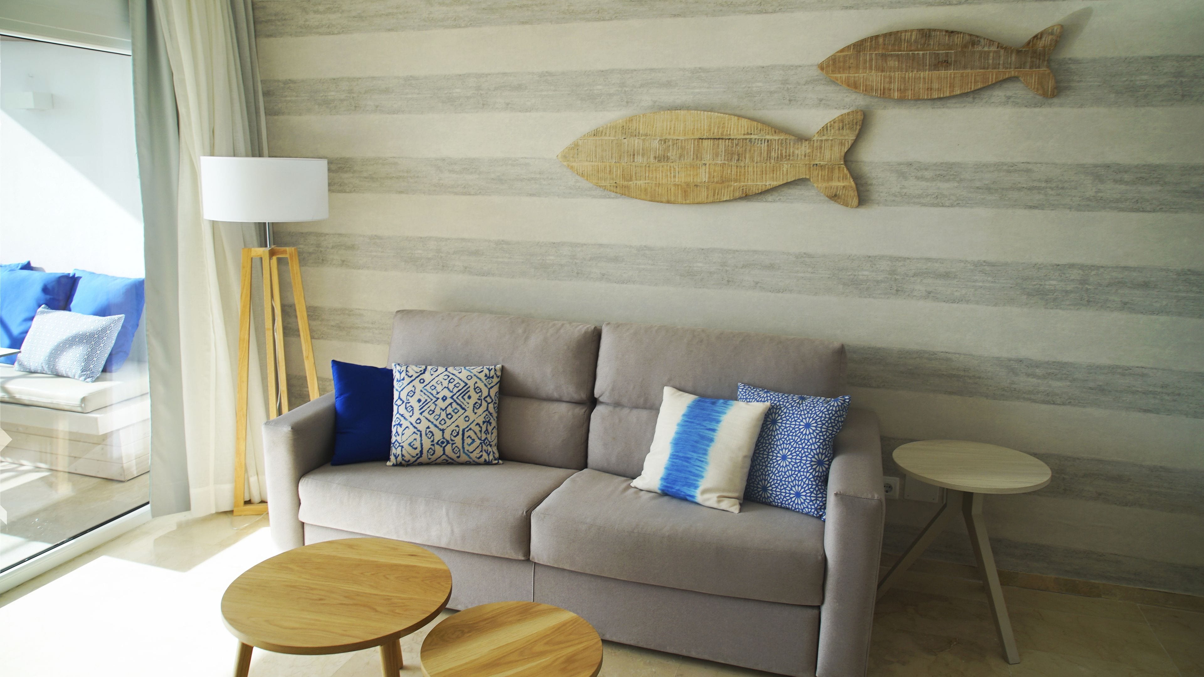 Una sala moderna con un sofá gris, cojines estampados, un adorno mural de pescado de madera, una lámpara de pie y dos mesitas de madera redondas.