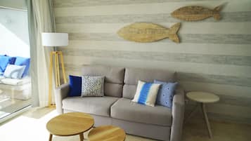 En moderne stue med en grå sofa, mønstrede puder, fiskemotiv på væggen, en gulvlampe og to runde træsideborde.