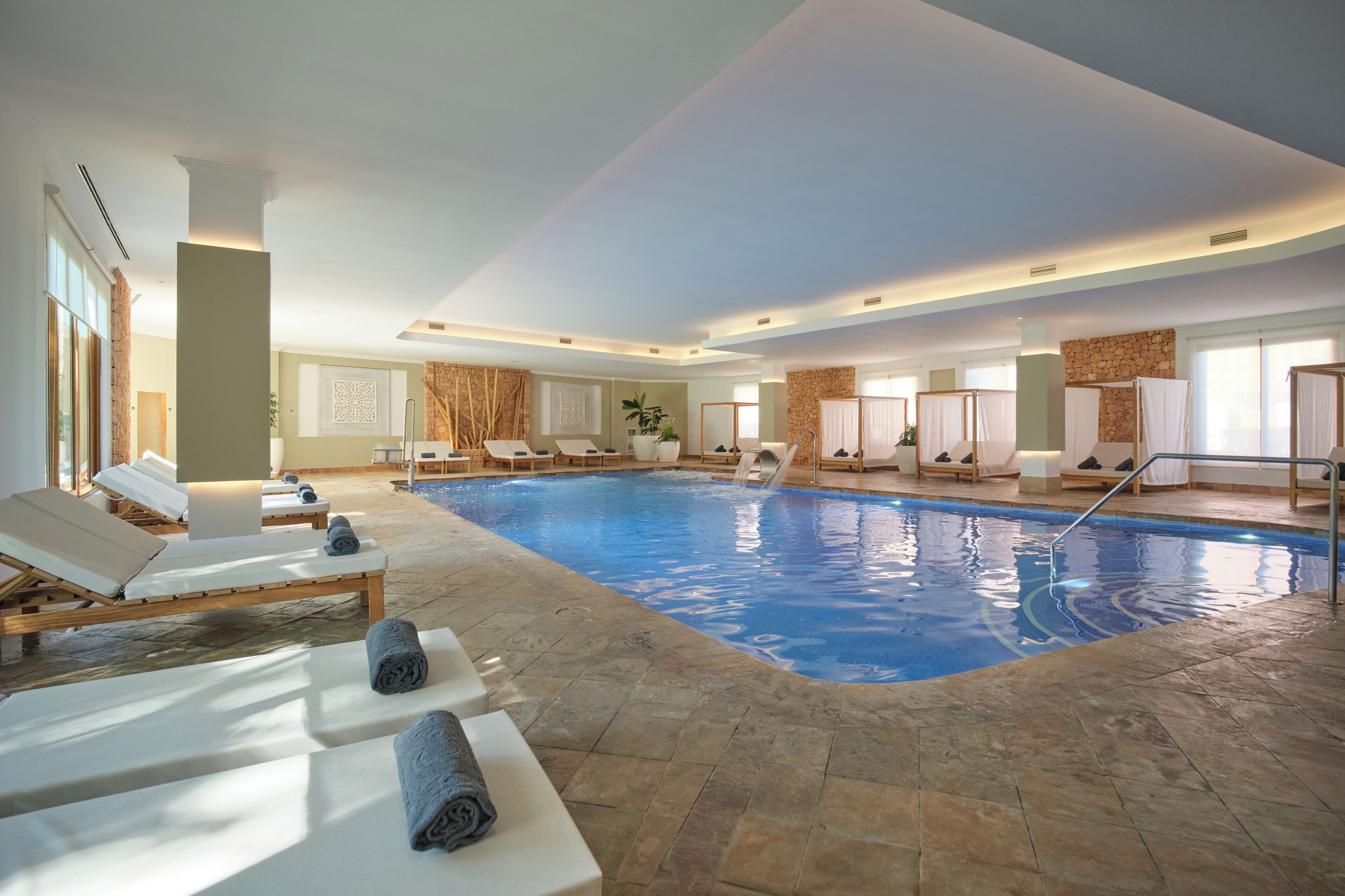 Una piscina interior con sillones de descanso y tumbonas, rodeada de paredes de piedra y ventanas grandes.