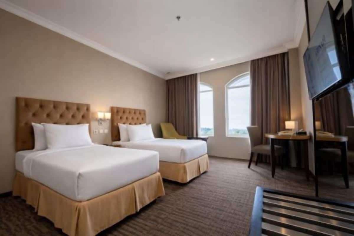 A hotel room with two beds, a TV, a desk, and a window with curtains.