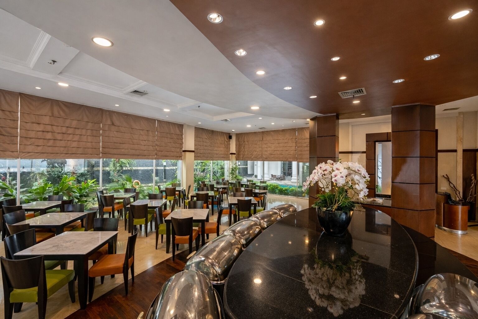 A spacious indoor dining area with multiple tables and chairs, large windows offering a view of greenery outside, and a central decorative plant arrangement.