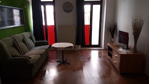 Sala de estar com sofá, uma mesa pequena, radiador, relógio e televisão sobre um móvel de madeira.