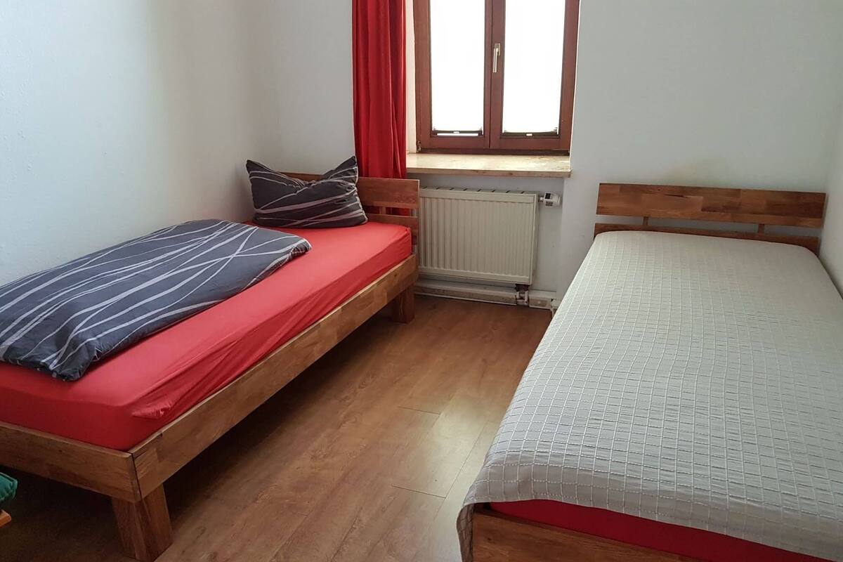 A small room with two wooden beds, one with a red mattress and the other with a white mattress.