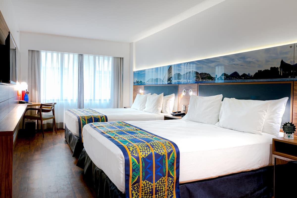 Quarto de hotel com duas camas, uma escrivaninha e uma janela grande.