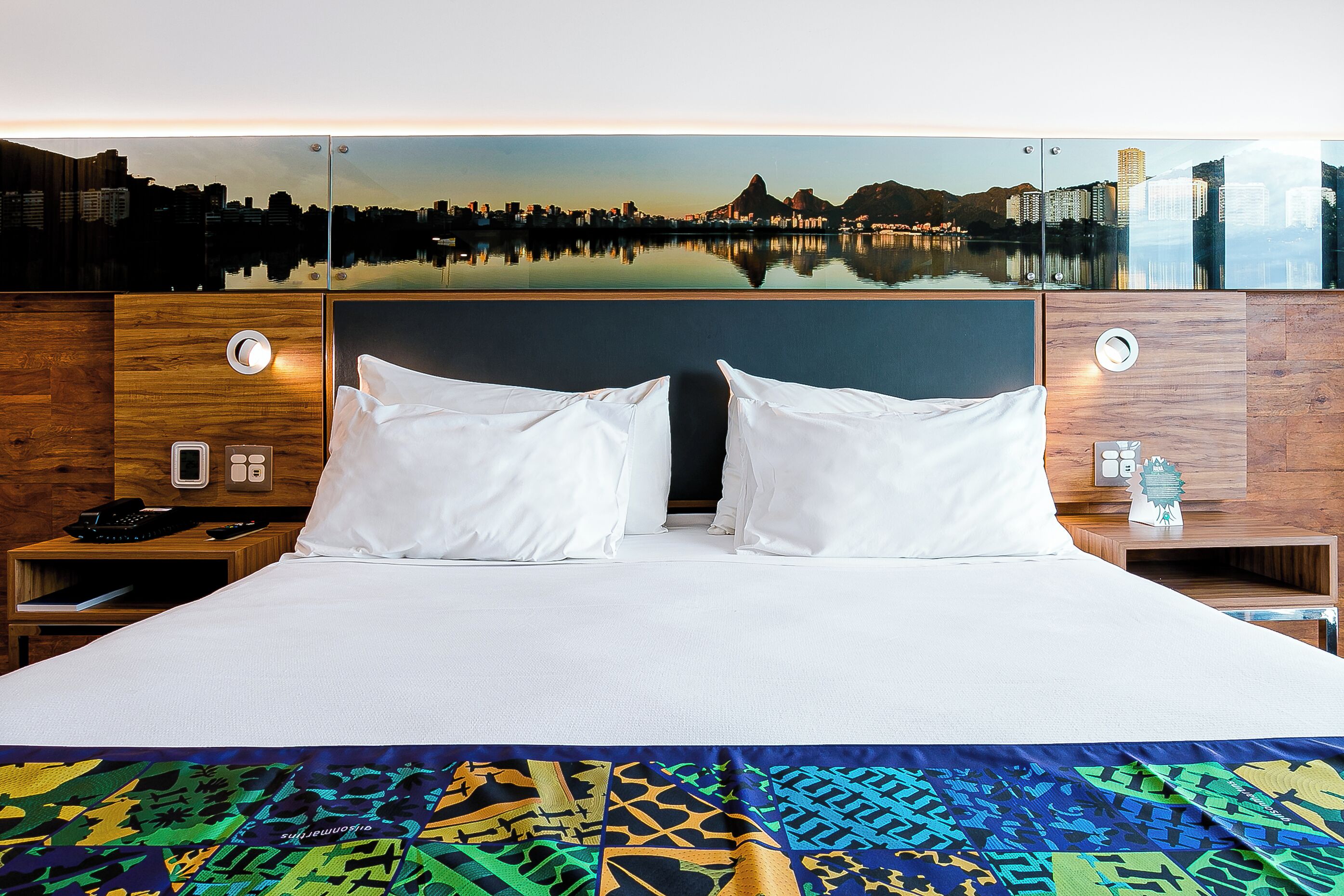 A hotel room with a large bed, two bedside tables, a nightstand, a cityscape view, and a wooden headboard.