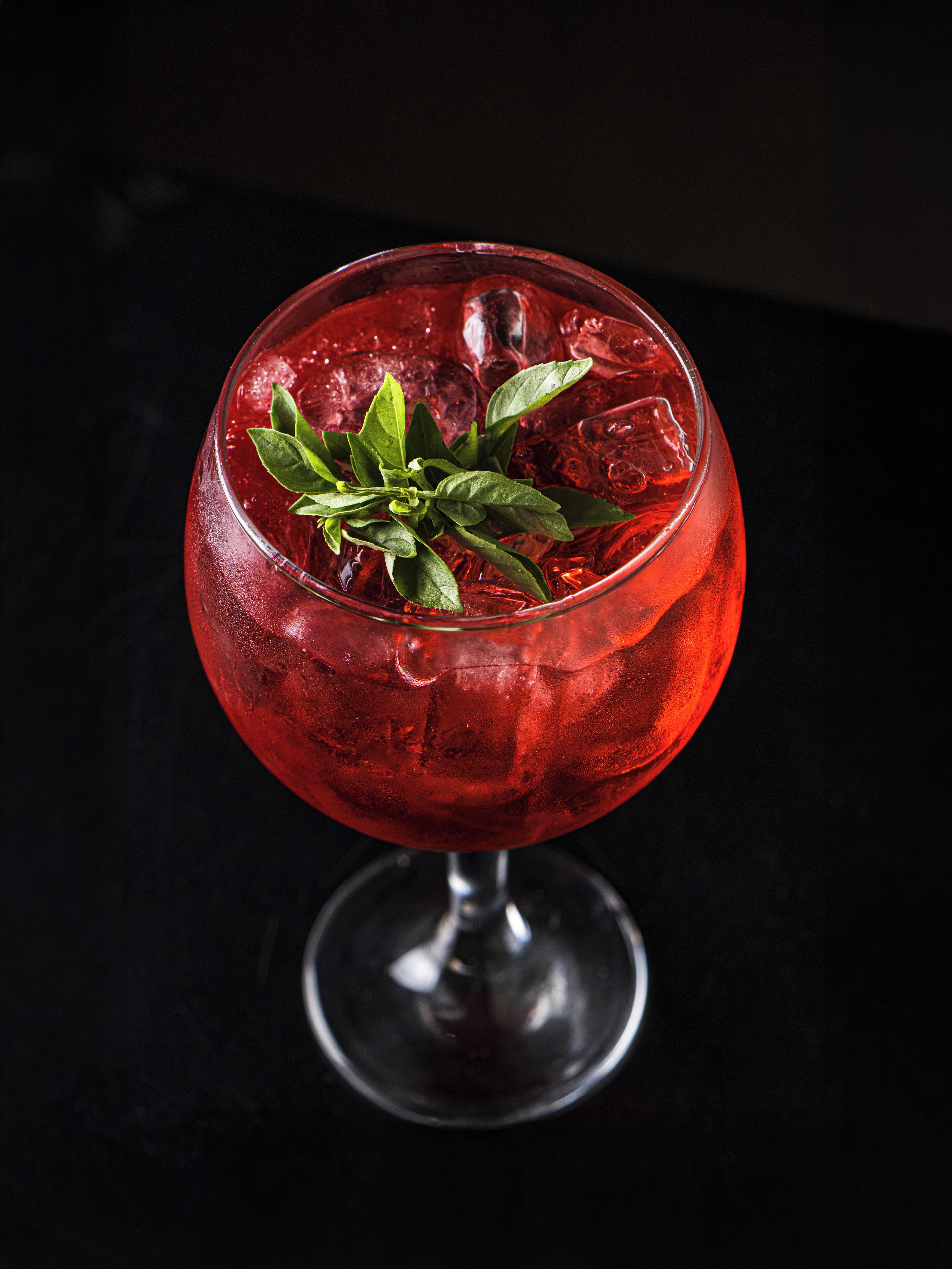 Un vaso de cóctel con una bebida roja, cubitos de hielo y una guarnición sobre una superficie oscura.