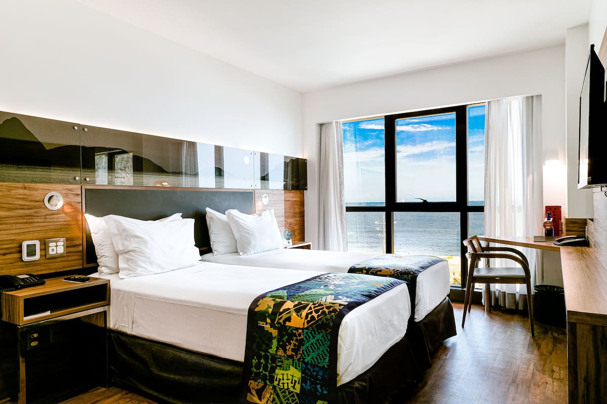 Quarto de hotel com duas camas, uma escrivaninha, uma cadeira, televisão e uma janela grande com vista para o mar.