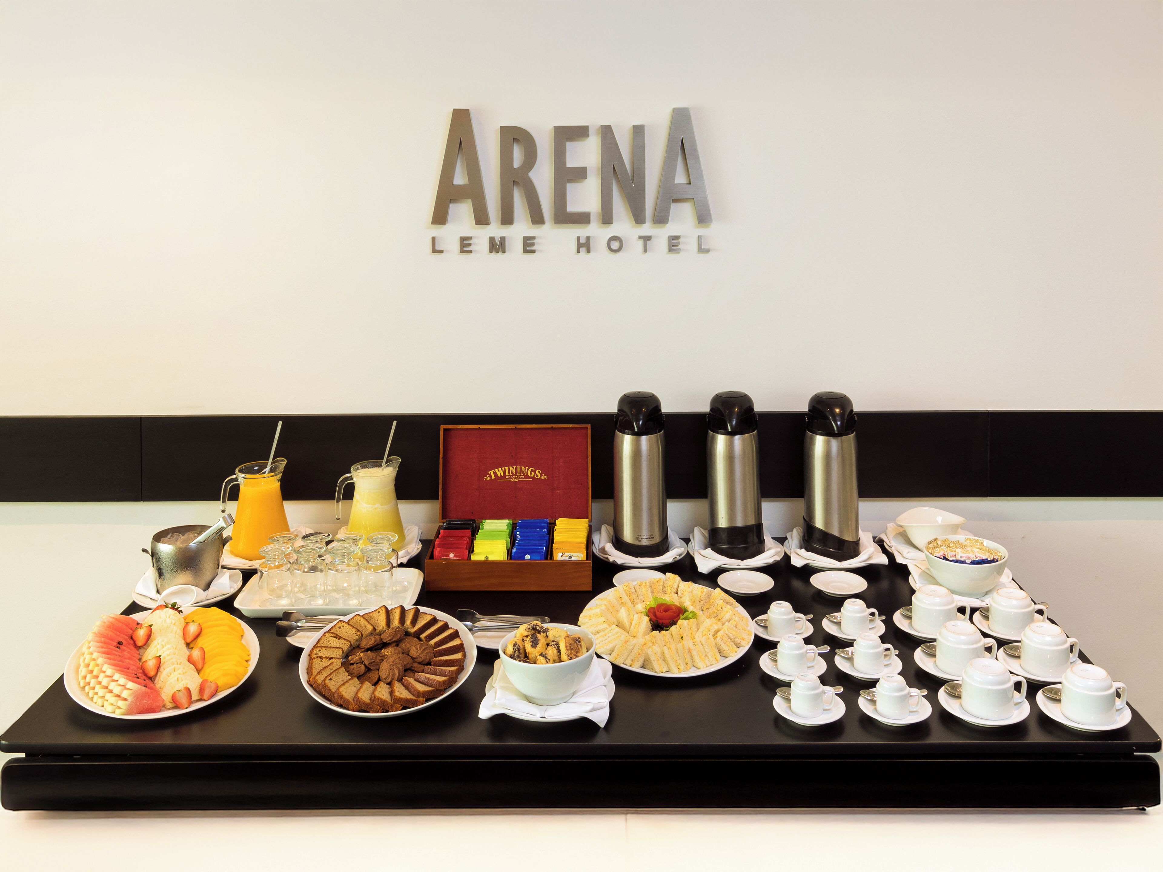 A breakfast buffet setup at the Arena Leme Hotel, featuring a variety of fruits, pastries, and beverages.
