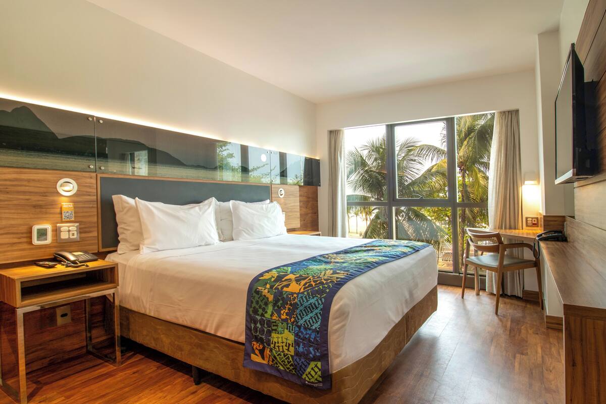 Quarto de hotel com cama, mesa de cabeceira, abajur e vista para palmeiras através de janelas amplas.