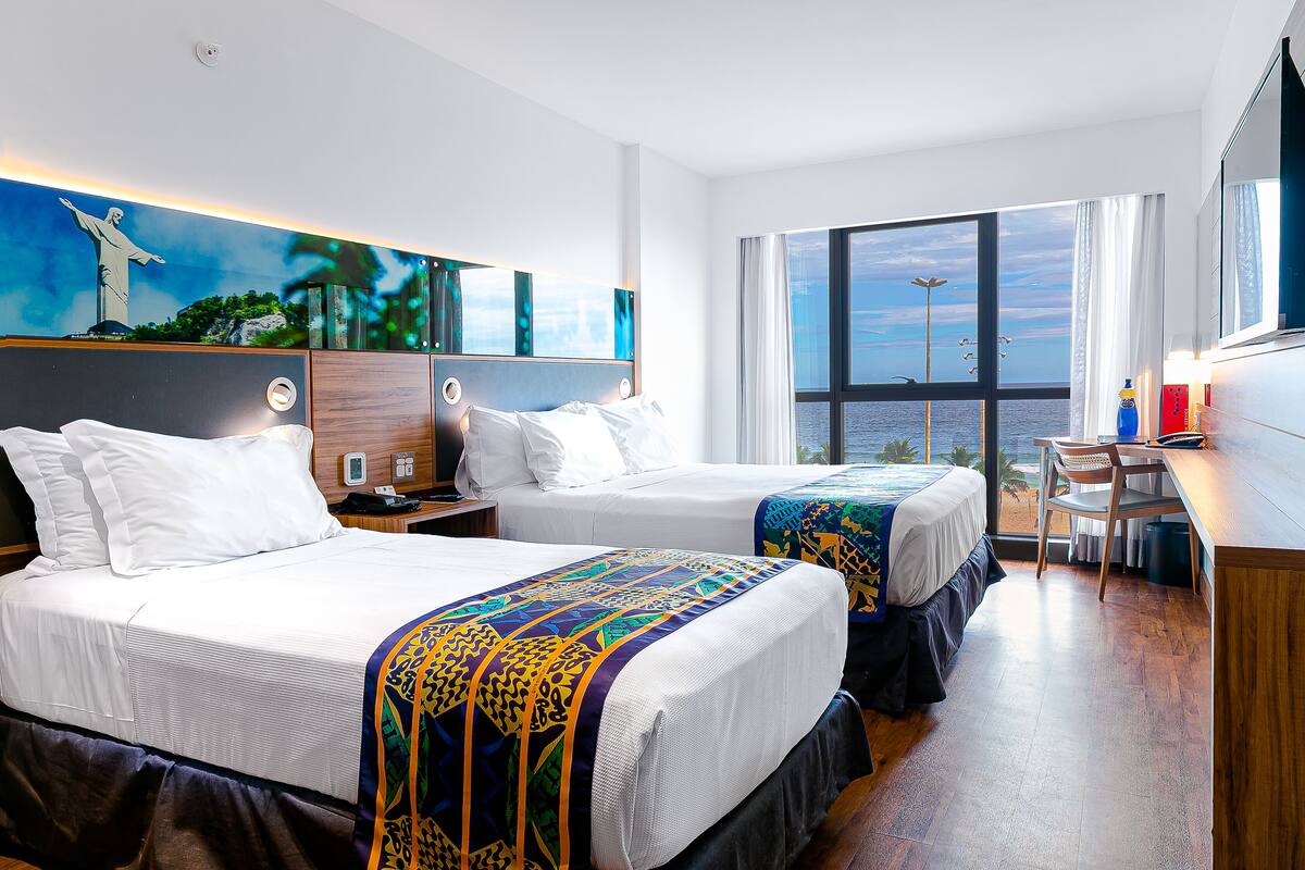 Quarto de hotel com duas camas, uma escrivaninha e uma janela grande com vista para o mar.