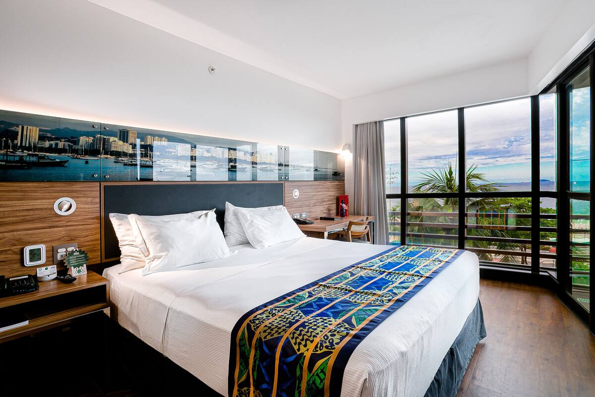Quarto de hotel com uma cama grande, uma escrivaninha e vista para a paisagem urbana através de uma parede de vidro.
