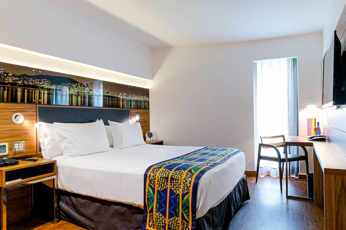 Quarto de hotel com cama, mesa de cabeceira, escrivaninha, cadeira e vista para a cidade pela janela.