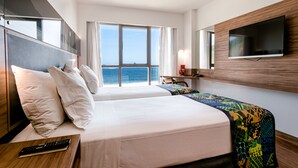 Ein Hotelzimmer mit zwei Betten, einem an der Wand montierten Fernseher, einem Schreibtisch mit Stuhl und einem großen Fenster mit Meerblick.