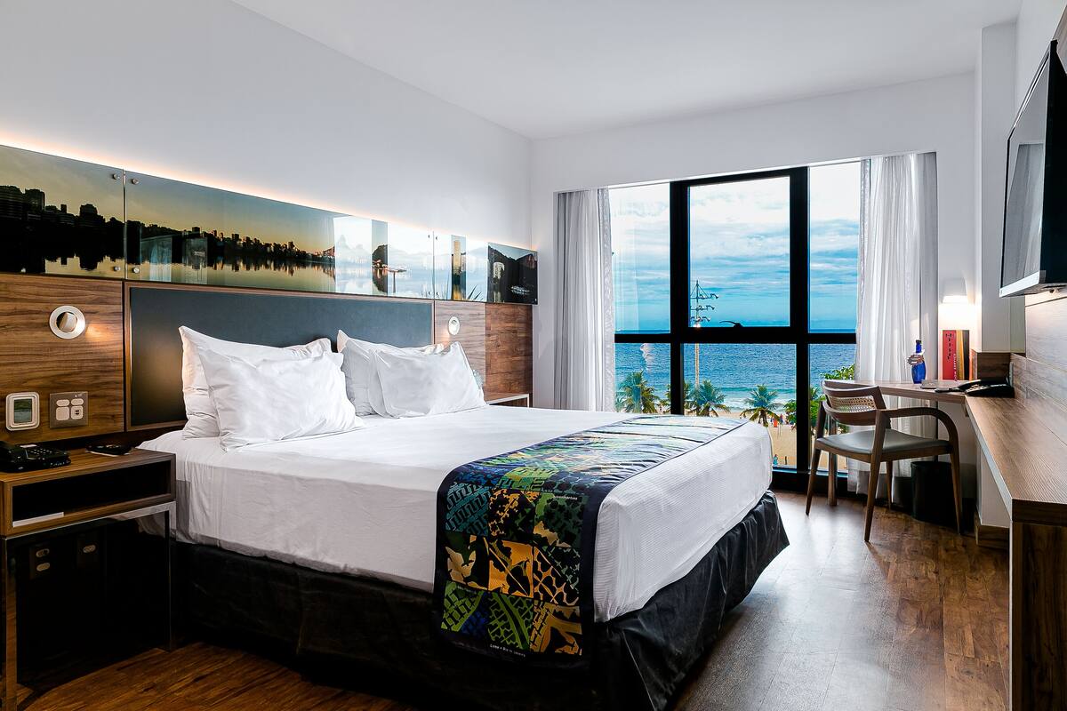 Quarto de hotel moderno com uma cama grande, uma escrivaninha e vista para o oceano.