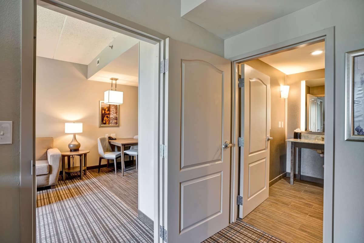 A hotel hallway with a carpeted floor, a dining area with a table and chairs, and a bathroom with a sink and mirror.