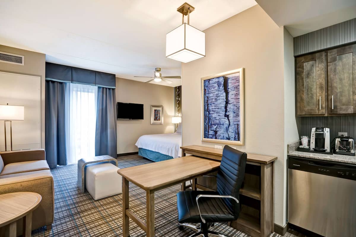 A hotel room with a bed, a desk, a chair, a sofa, a small table, a ceiling fan, a painting, and a kitchenette.