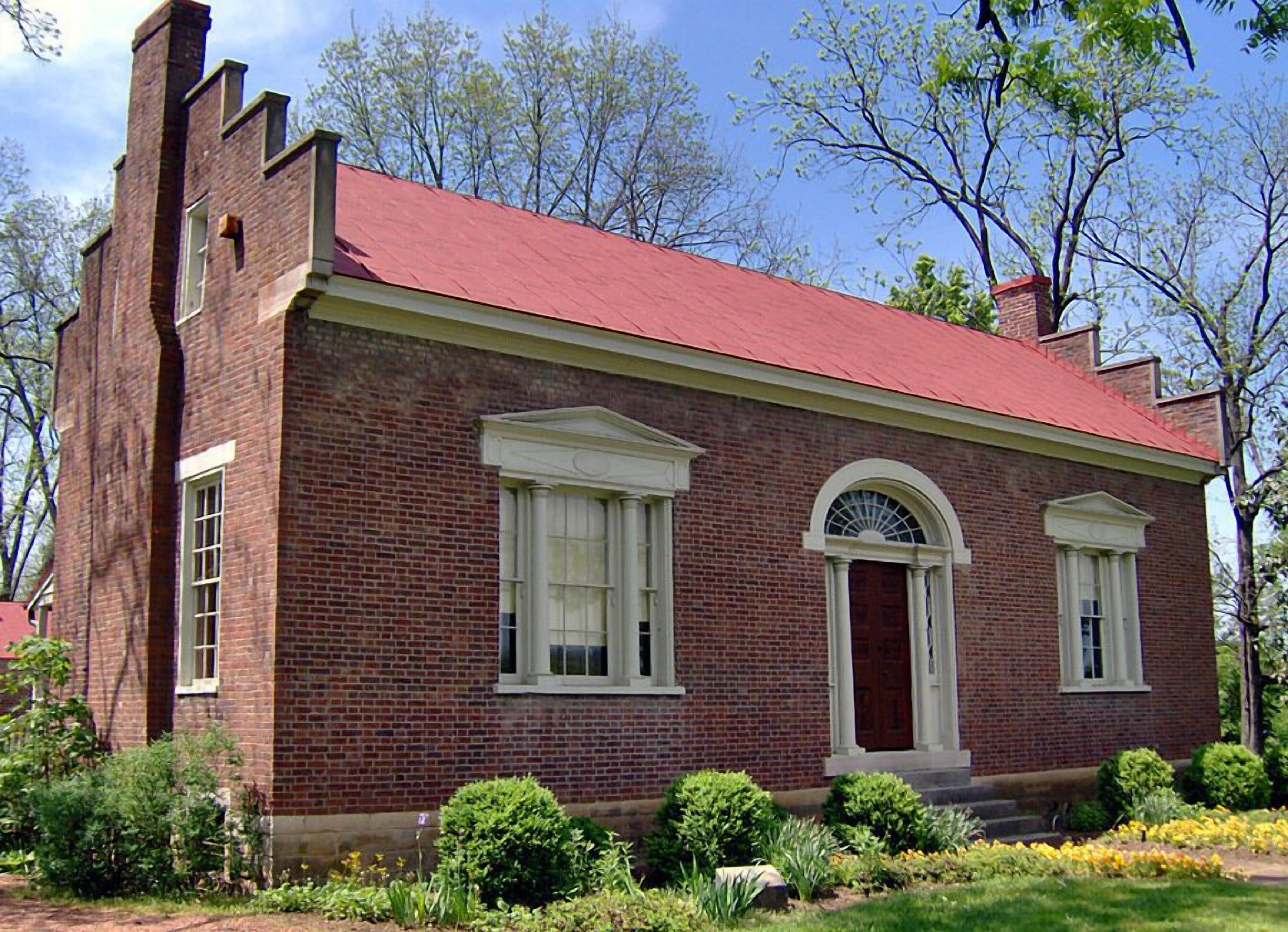 A brick building with a red roof, two large windows with white frames, and a central arched doorway.