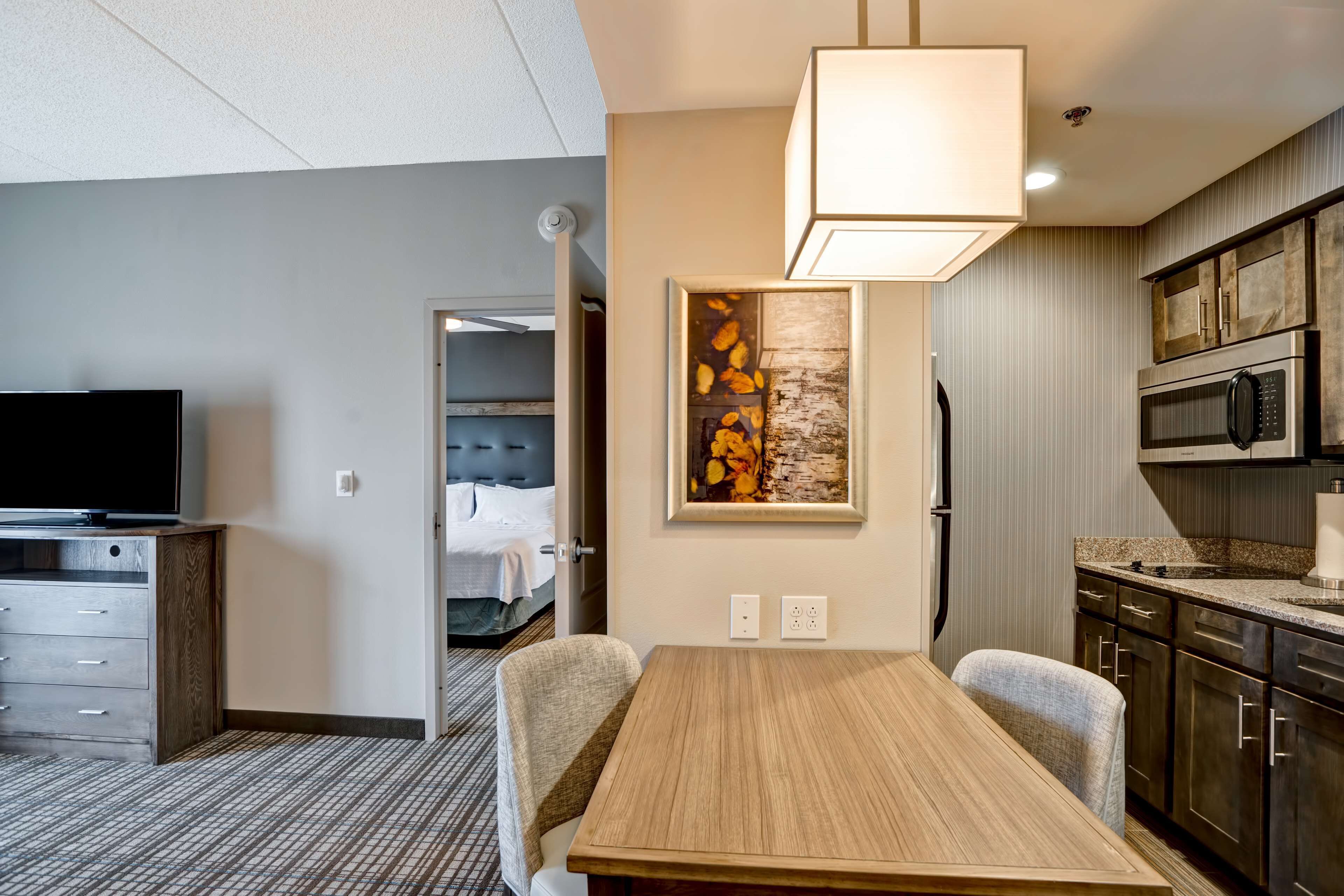 A hotel room with a dining table, chairs, a TV, a painting, and a kitchenette.