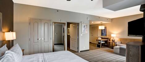 A hotel room with a large bed, a ceiling fan, a desk with a chair, a television, and a small seating area.
