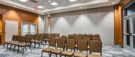 A conference room with rows of chairs and tables, a wooden wall, a large window, and a ceiling with recessed lighting.