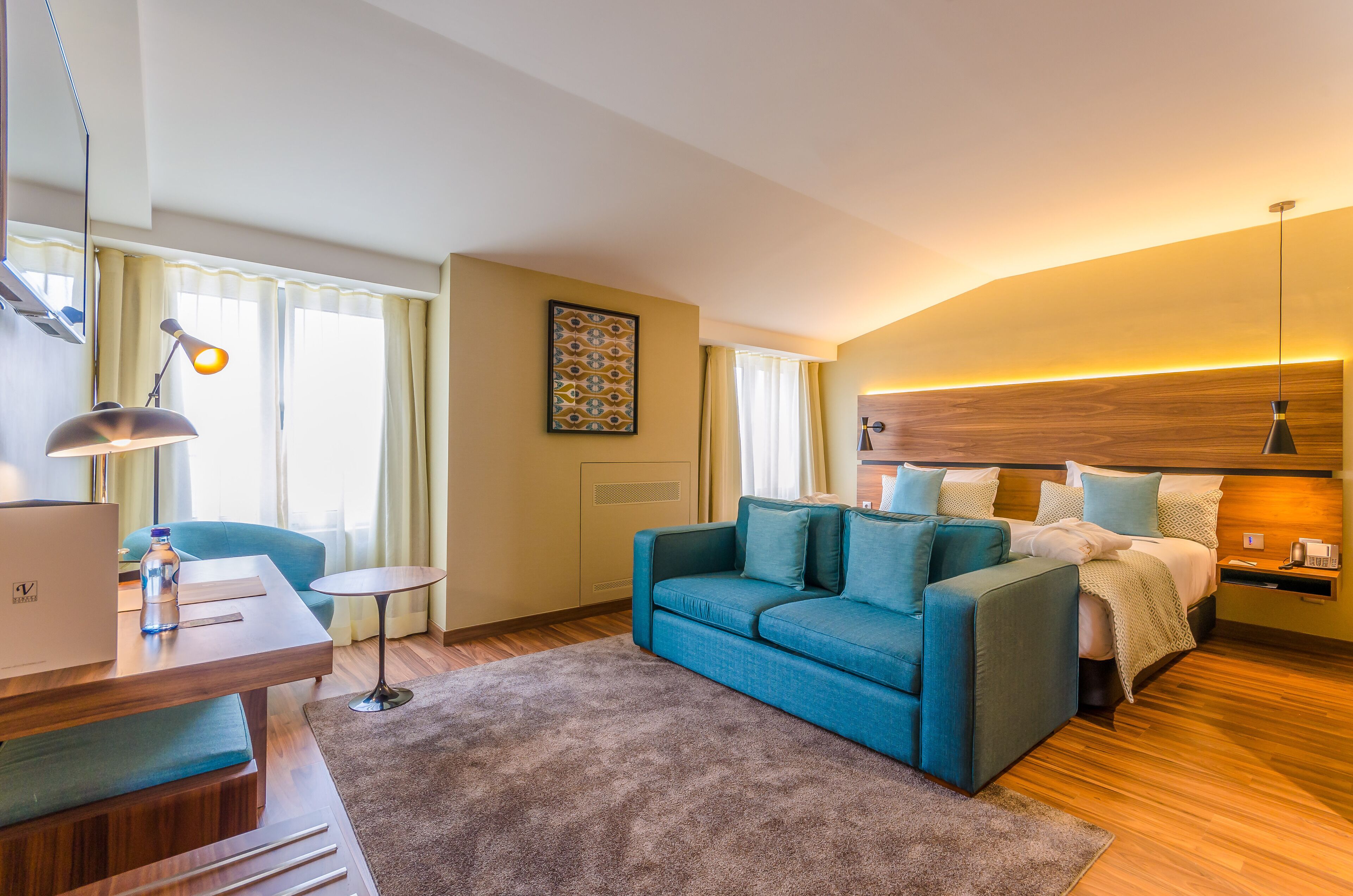 A modern hotel room with a blue sofa, a bed with white linens, a desk with a laptop, and a small round table.