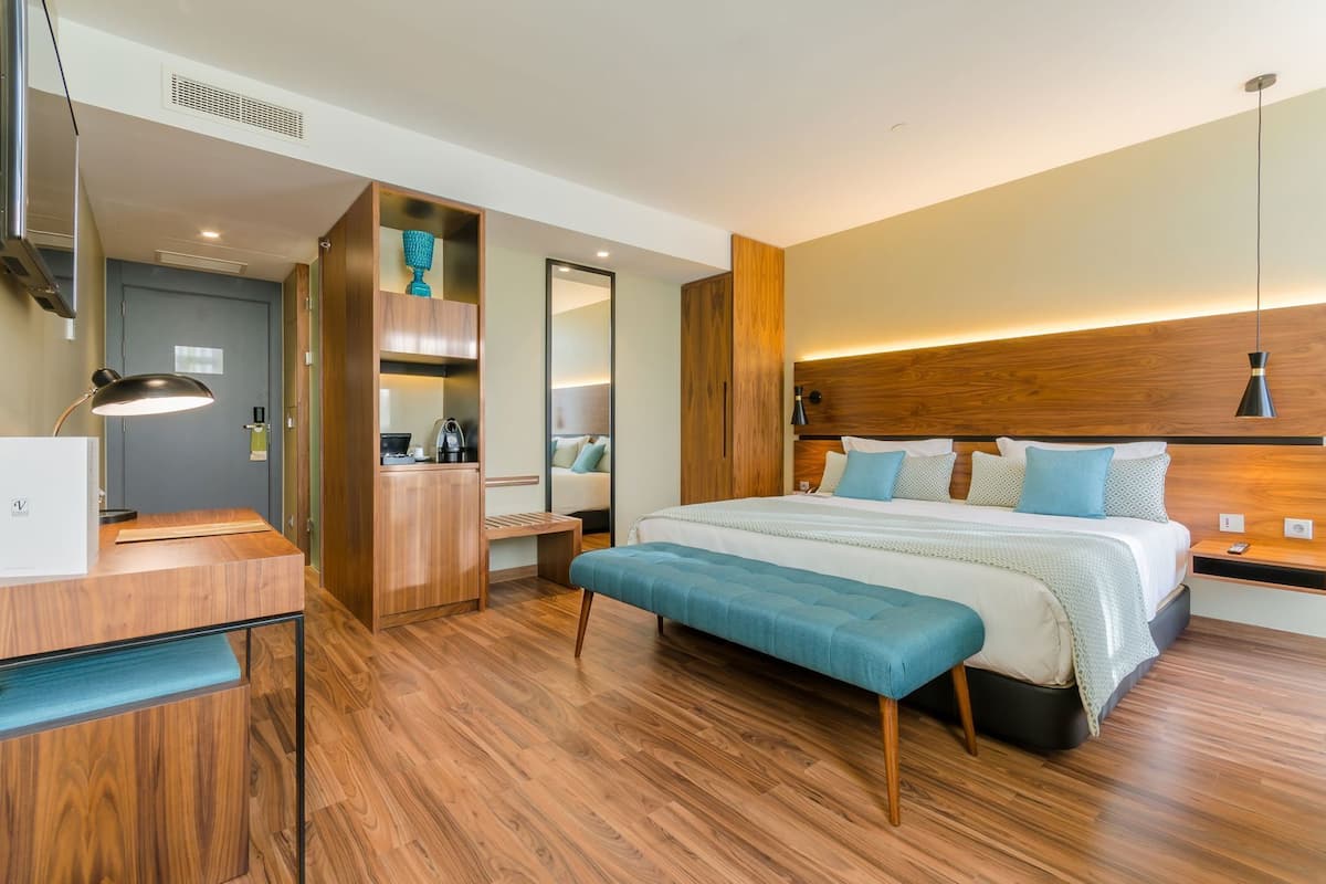 A modern hotel room with a wooden floor, a bed with multiple pillows, a desk with a lamp, a wardrobe, and a mirror.