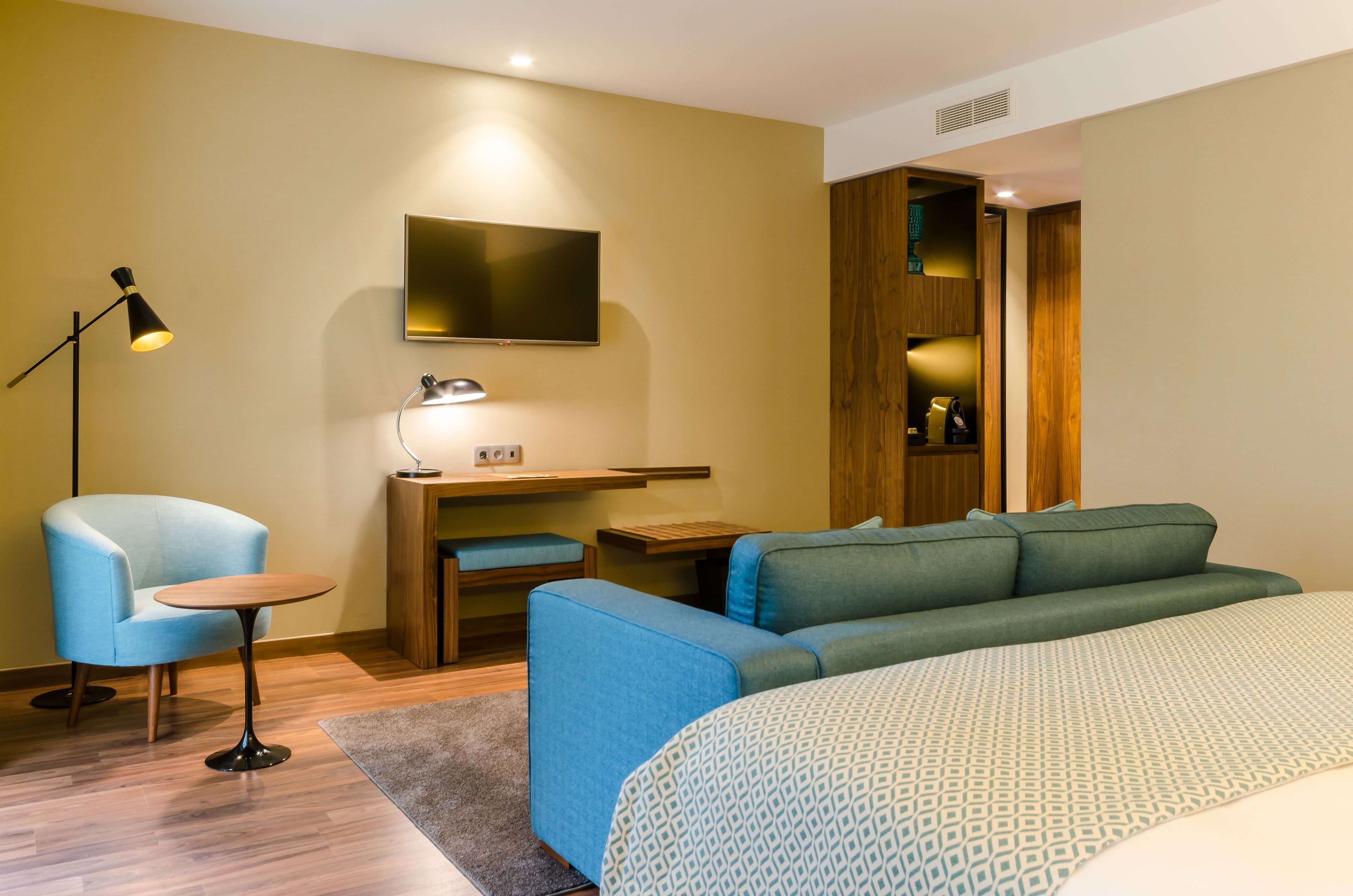 A modern hotel room with a bed, a blue sofa, a desk with a chair, a TV mounted on the wall, and a lamp.
