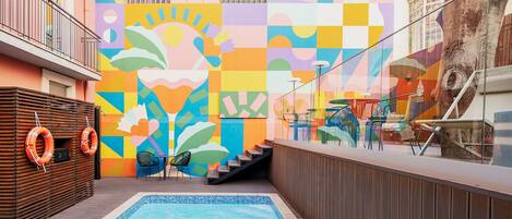 Zona de piscina con un mural colorido, una terraza de madera y mobiliario exterior.
