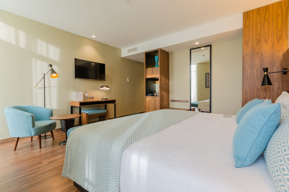 A modern hotel room with a large bed, a desk, a chair, a TV, and a wardrobe.