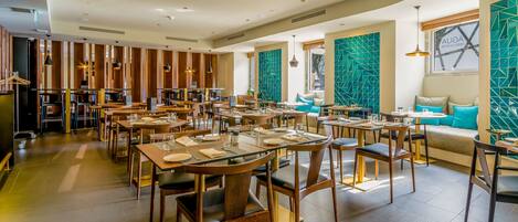 A spacious restaurant with multiple dining tables and chairs, a high ceiling with recessed lighting, and a large window with a geometric wall design.