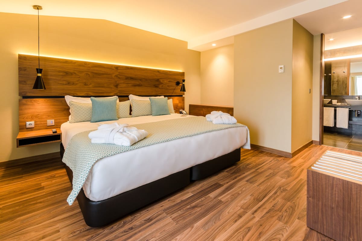 A modern hotel room with a large bed, wooden headboard, bedside tables, and a built-in bench.