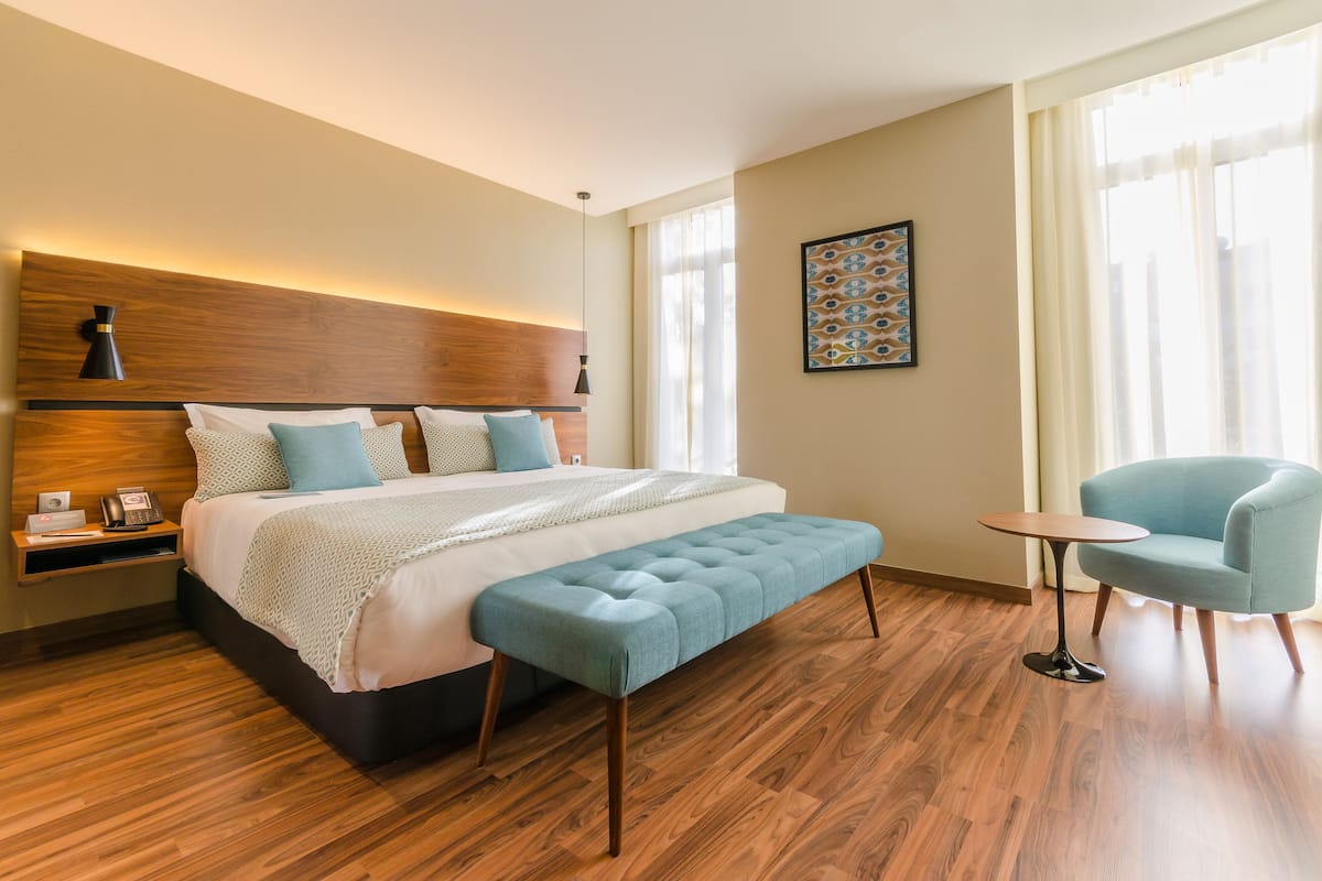 A modern hotel room with a large bed, a bedside table, a small round table, a blue armchair, and a wooden floor.
