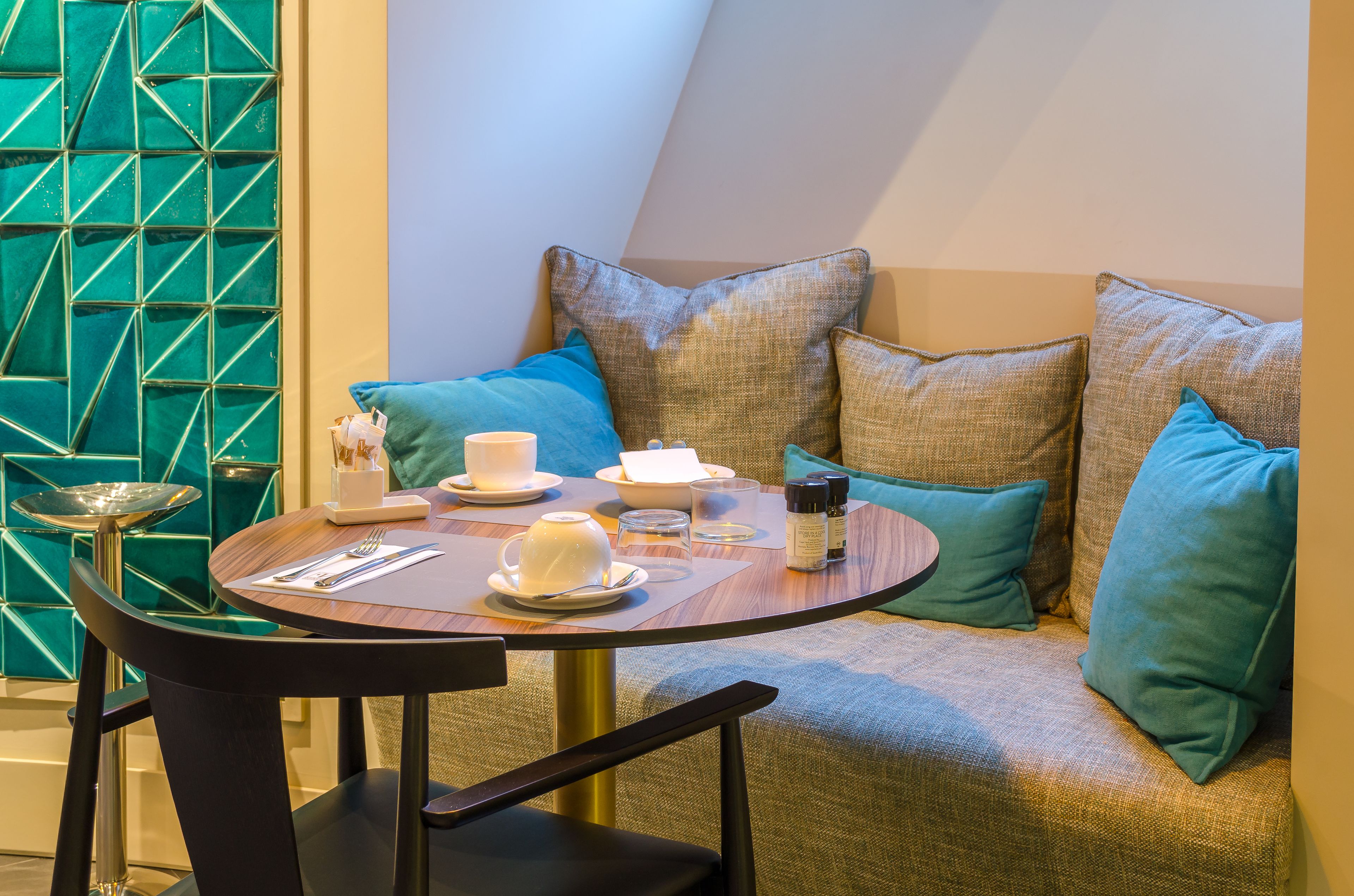 Un rincón acogedor con una mesa redonda preparada para el té, una pared verde con estampado y un sofá cómodo con cojines azul verdosos.