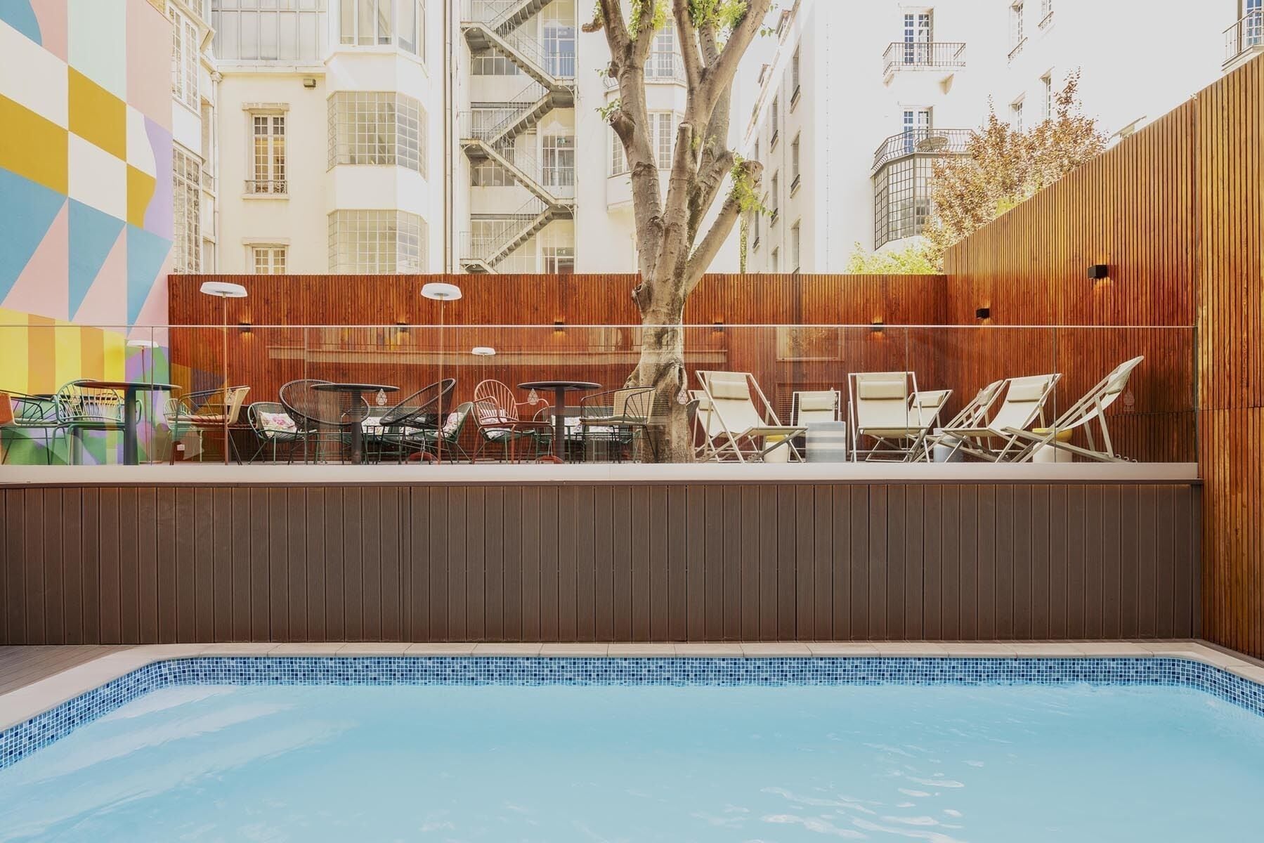 Un espace extérieur avec piscine, comprenant des chaises longues et des tables, entouré d’une clôture en bois et d’un mur aux couleurs vives.