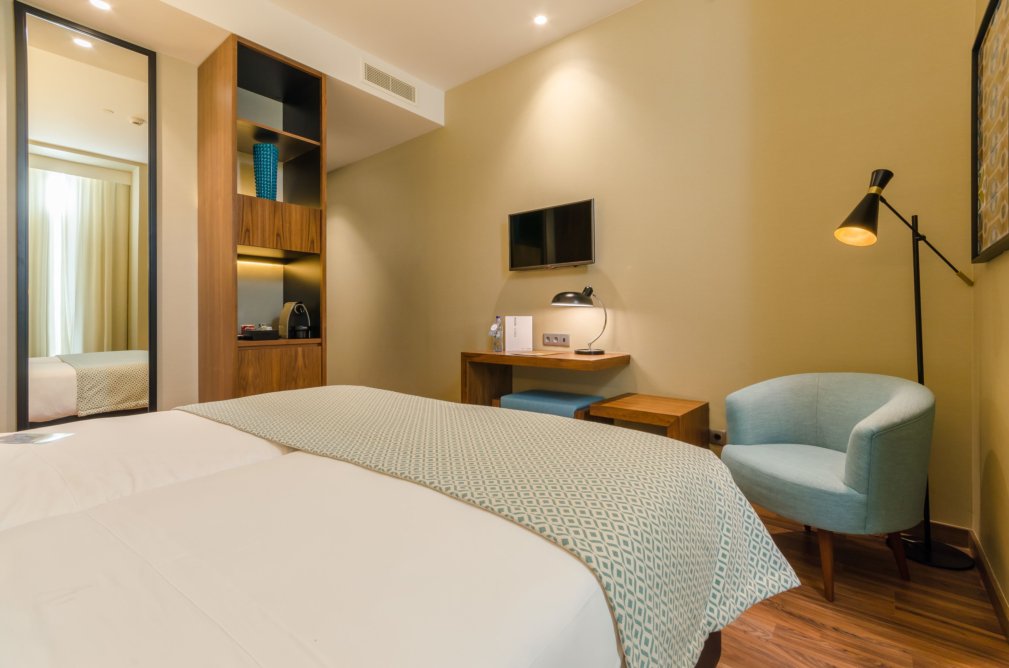 A hotel room with a large bed, a desk with a chair, a TV, and a floor lamp.