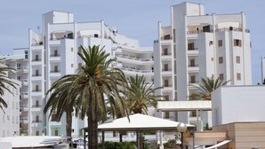 Un bâtiment moderne de couleur blanche, doté de nombreux balcons, de palmiers et d’un espace extérieur aménagé avec des sièges, protégé par un auvent.