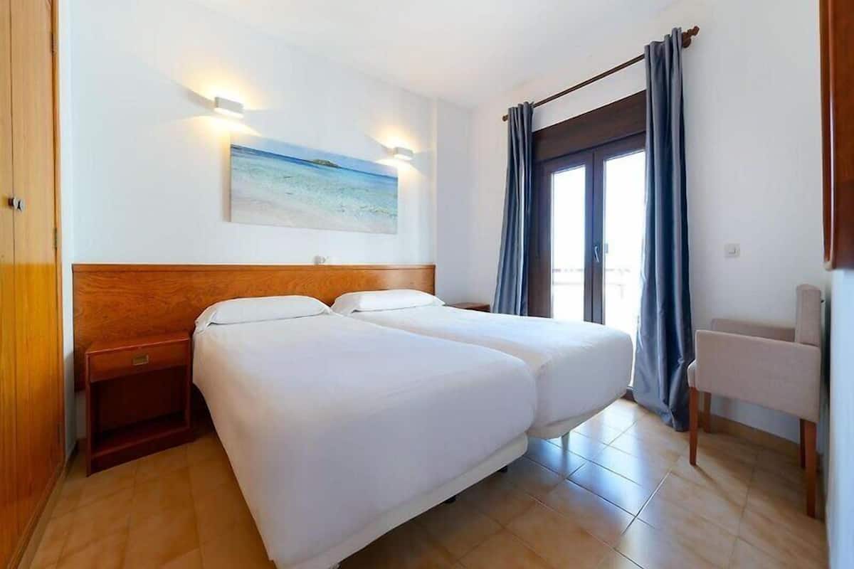 Habitación de hotel con dos camas, cabecera de madera, mesita de noche, una silla, un cuadro y vistas al exterior.