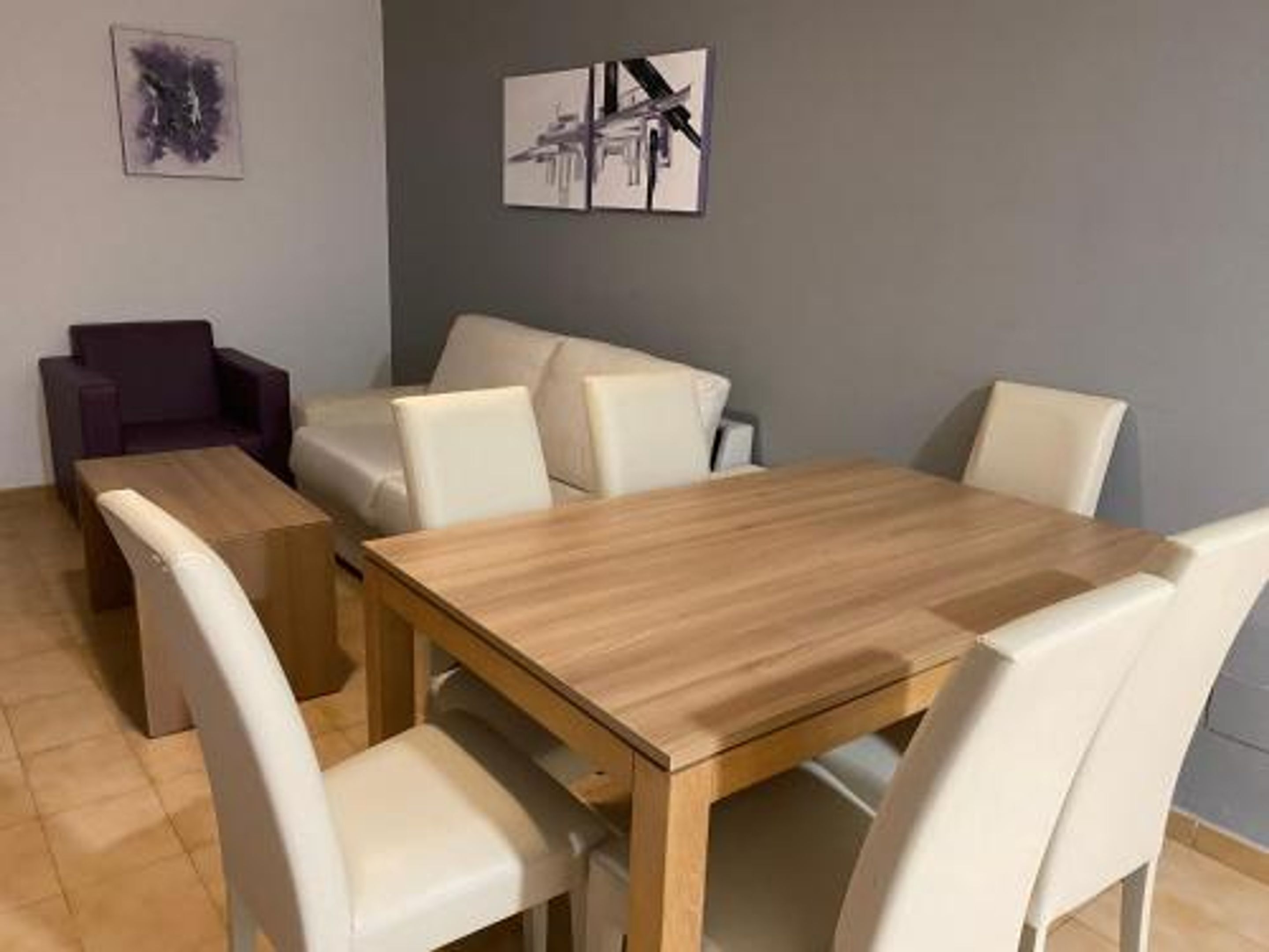 Un área de comedor con una mesa de madera y sillas blancas, un sofá morado y dos obras de arte enmarcadas en la pared.