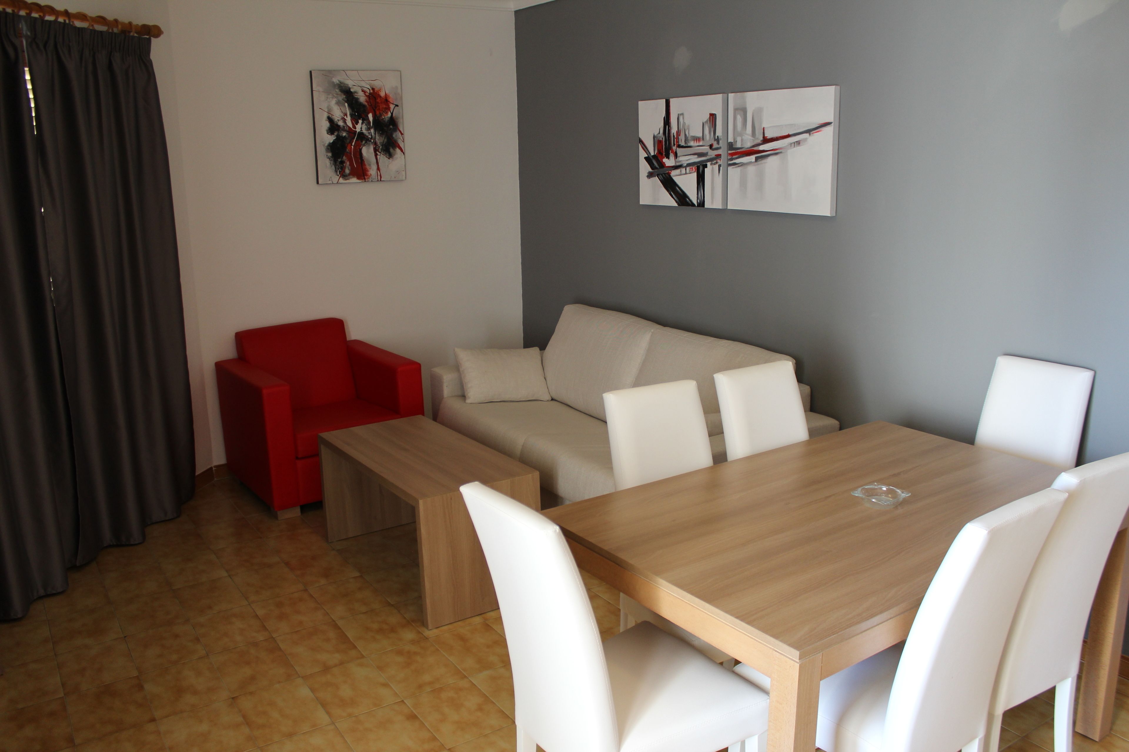 Un soggiorno moderno con un tavolo da pranzo in legno, sedie bianche, una poltrona rossa e un divano beige. Alle pareti sono esposti dipinti astratti.