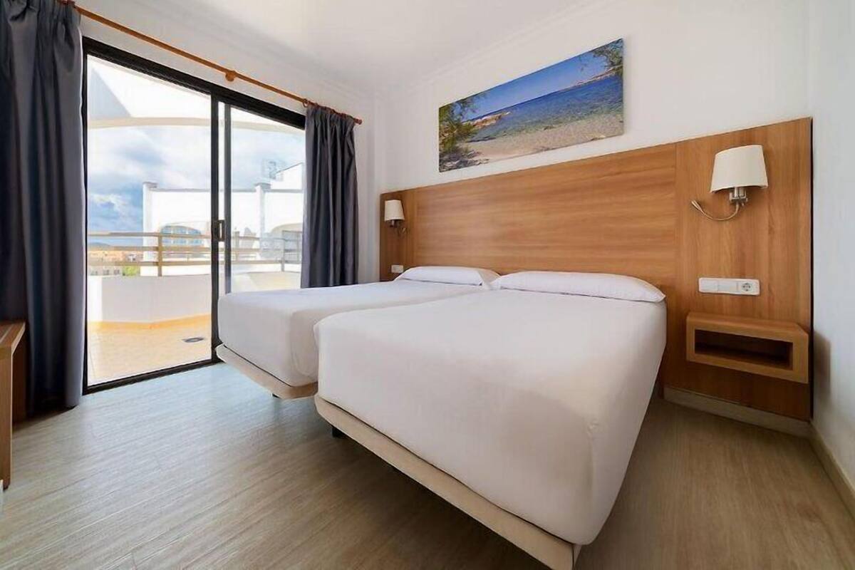 Habitación de hotel con dos camas, un ventanal con cortinas, un cuadro de una playa y un cabecero de madera.