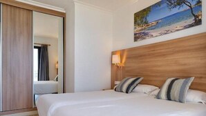 Une chambre d’hôtel avec un lit, une tête de lit en bois, un tableau représentant une plage et une fenêtre avec des rideaux.