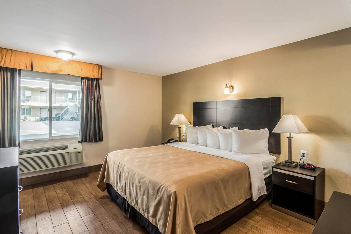A hotel room with a large bed, two bedside tables with lamps, a window with curtains, and a view of the outside.