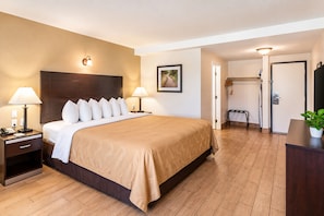 A hotel room with a bed, bedside lamps, a nightstand, a desk, a chair, a potted plant, and a door. - Quality Inn & Suites near Downtown Bakersfield (Bakersfield)