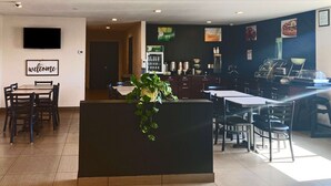 Un vestíbulo de hotel con una recepción, un cartel de 'welcome', una máquina de café y un área de descanso con mesas y sillas.