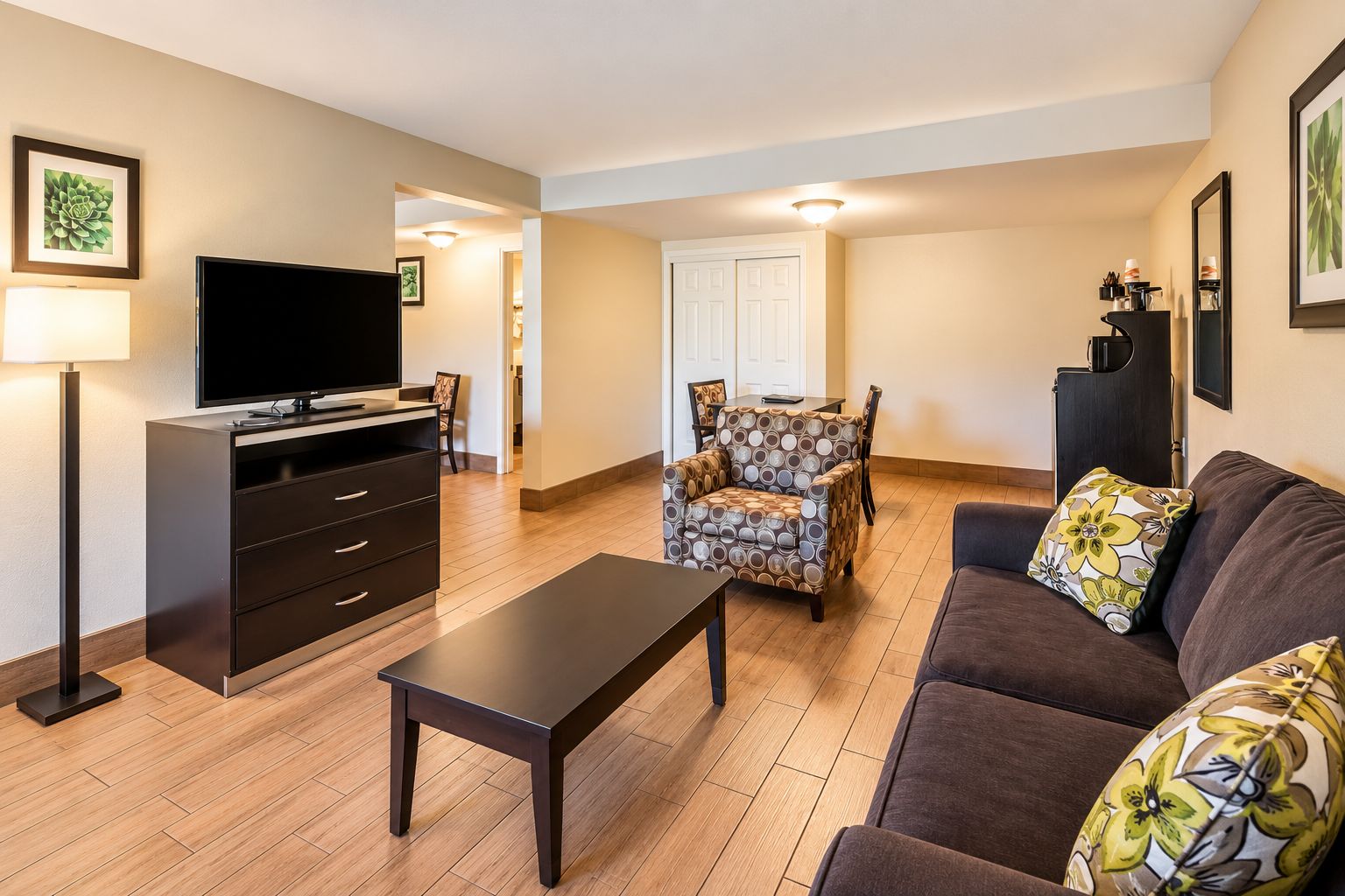A living room with a flat-screen TV, a dark wooden coffee table, a patterned armchair, a brown sofa with floral cushions, and a black cabinet.