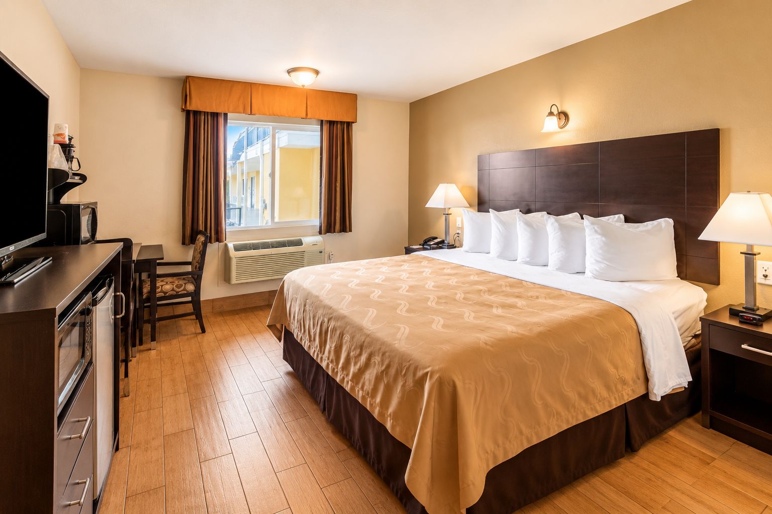 Una habitación de hotel con una cama grande, lámparas de noche, un escritorio con televisor y una ventana con cortinas.
