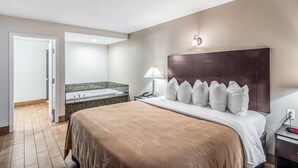 Habitación de hotel con una cama grande, cabecera, mesita de noche, lámpara, teléfono y baño con bañera y lavabo.