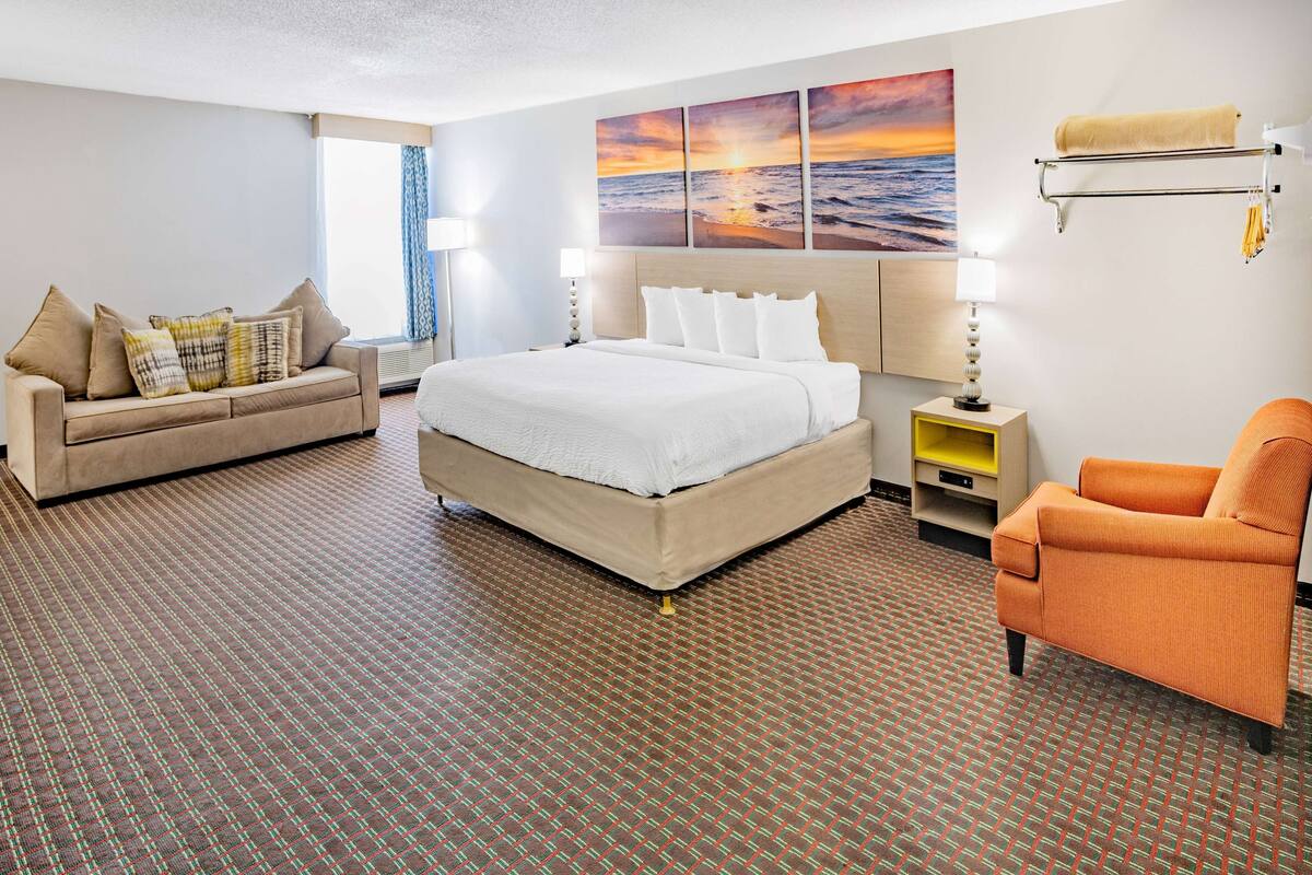 A hotel room with a large bed, a sofa, an orange armchair, a wall-mounted towel rack, and a sunset view.