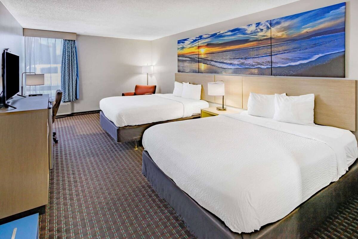 A hotel room with two beds, a desk, a TV, and a large painting of a sunset over the ocean.